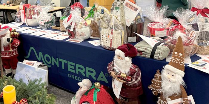 Photo of auction items including wrapped baskets and standing Santa Claus figures on a table with a blue Monterra Credit Union branded table drape.