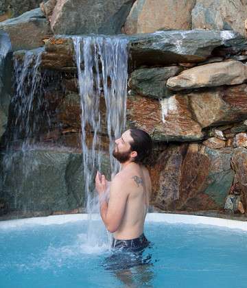 Image of a man in a pool, under a waterfall, with rock formations around him.